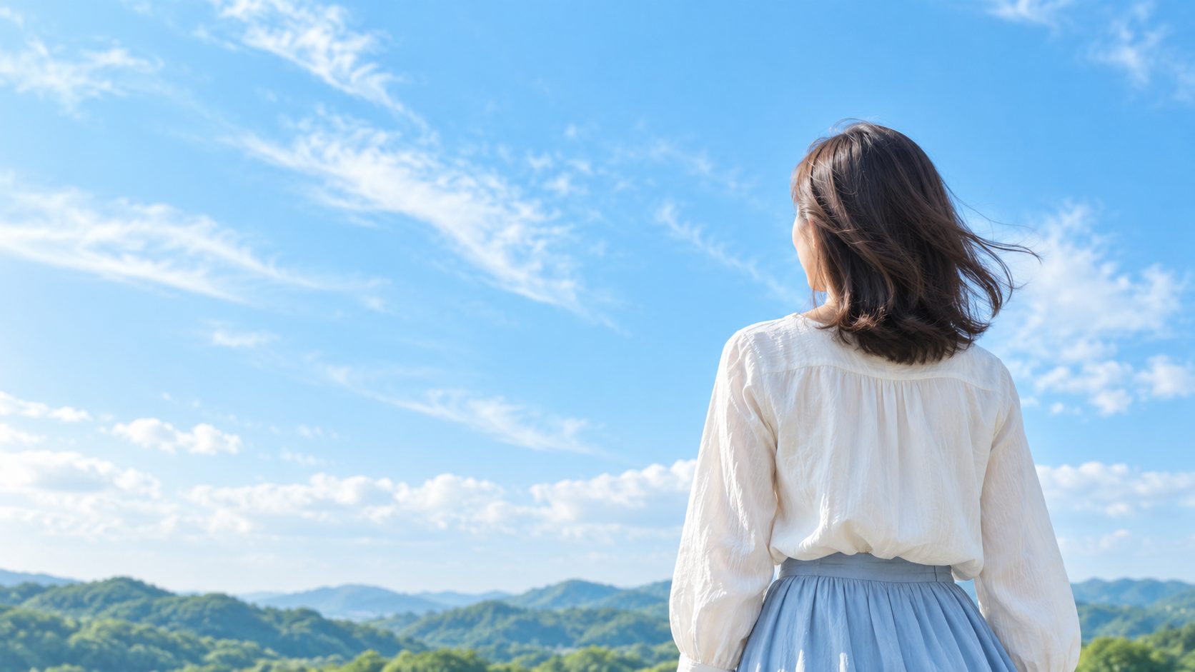 青空の下で、白いブラウスの女性が山並みを眺めているイメージ